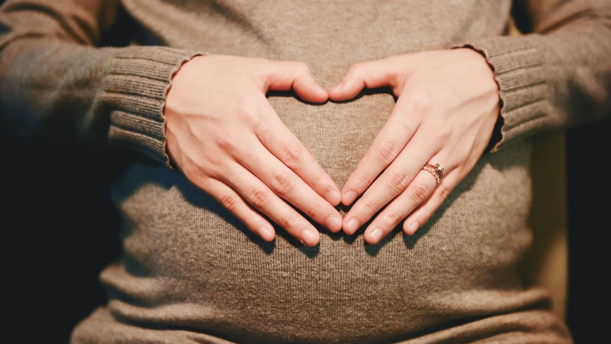 A person wearing a sweater forms a heart shape with their hands over their pregnant belly.